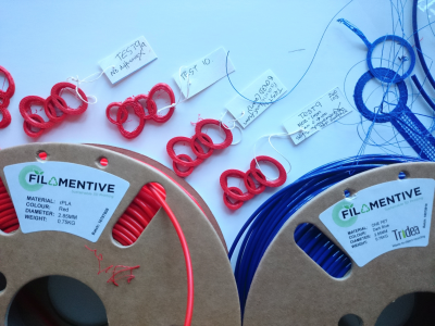 Image shows two reels of sustainable 3D printer filament in red and blue, plus 4 3D printed test pieces in red filament.