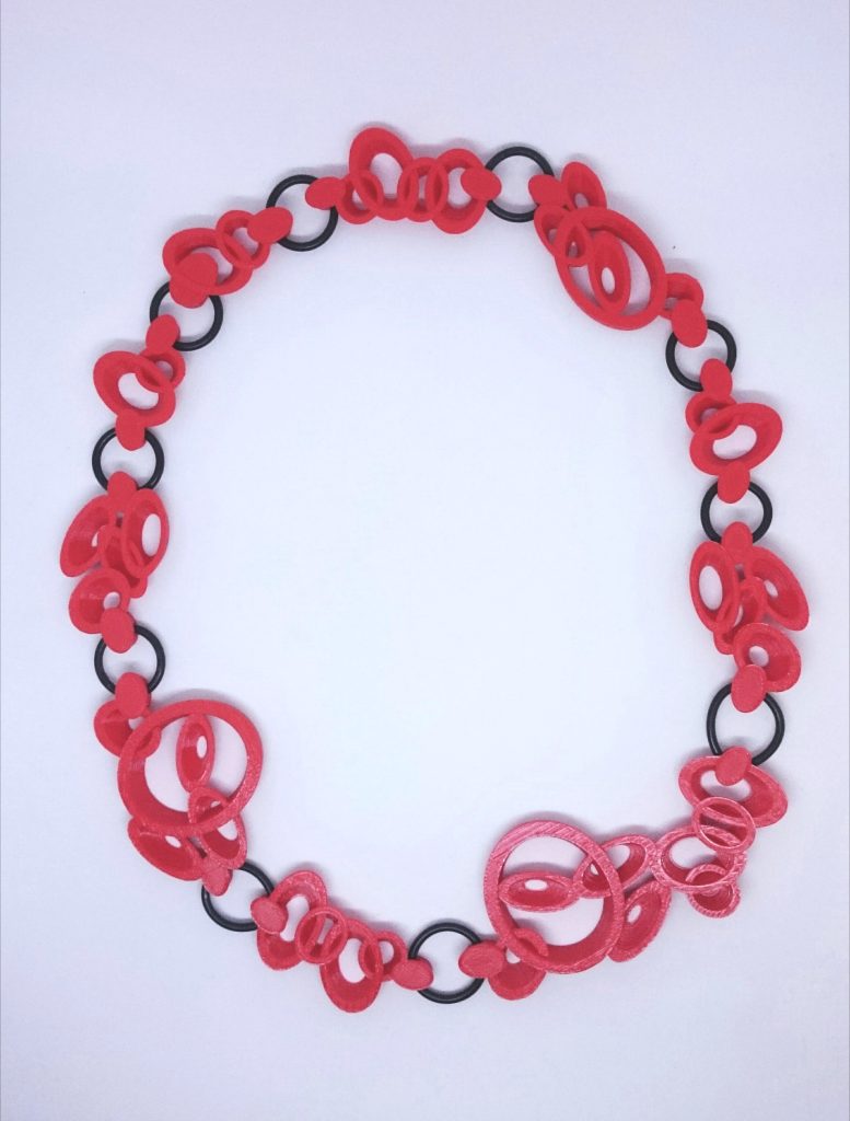This image shows a necklace made from 3D printed large red swirls joined wirh black O-rings. The inspiration for the Red Red necklace came via Ann Marie Shillito's lockdown project exploring 3D printing on fabric and other stuff as she had direct access to a desktop 3D printer and experimented with embedding materials during the 3D printing process. She loves O-rings for their potential, particularly as links as they are flexible and robust. These are left over from an earlier project and ideal for joining the units into a necklace during the 3D printing process. Just love the red and black combination.